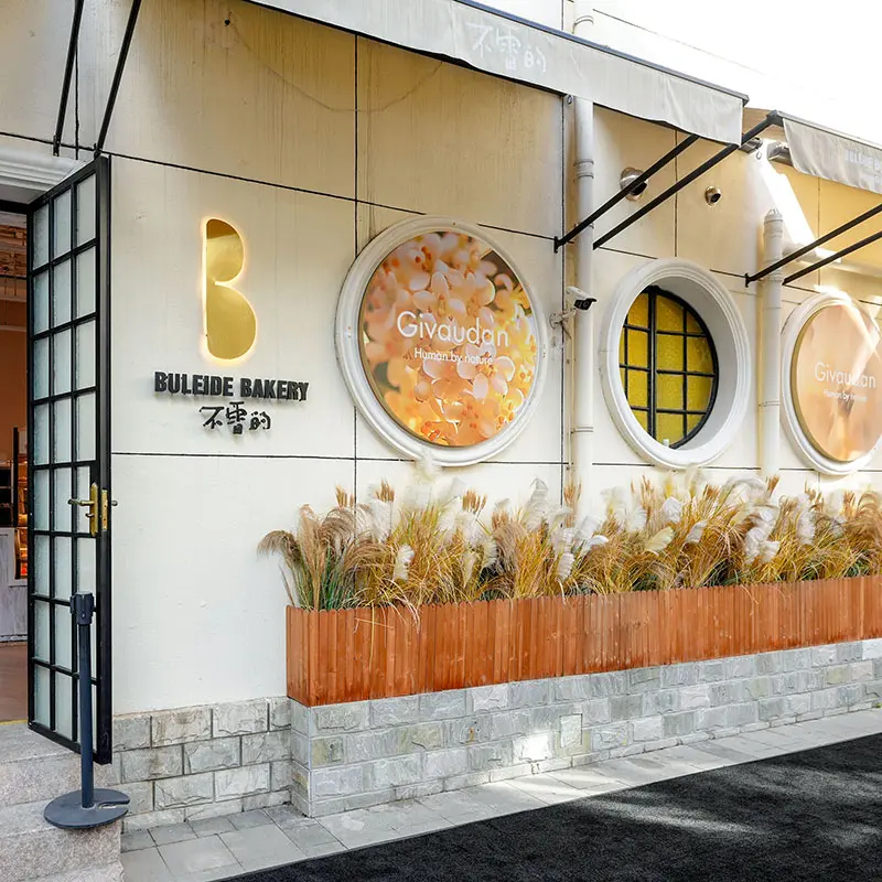 The entrance of Buleide Bakery features large glass doors and a stylish sign. The exterior is decorated with round, colourful displays and ornamental grasses in wooden planters, creating a welcoming atmosphere. Inside, glimpses of a well-designed interior are visible, hinting at a modern space.