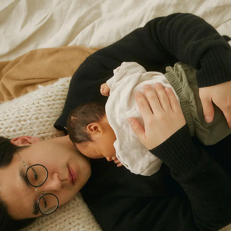 A man wearing glasses lies on a light-coloured blanket, softly resting with a baby nestled against his chest. The baby is dressed in a light sweater and green trousers, and the scene conveys a sense of warmth and intimacy.