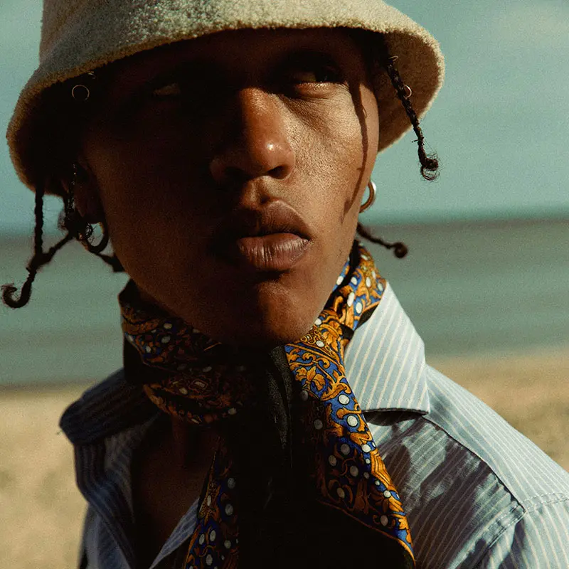 A person stands on a beach with a blurred ocean background, wearing a light blue striped shirt and a patterned scarf. The warm sunlight creates a relaxed, contemplative mood.