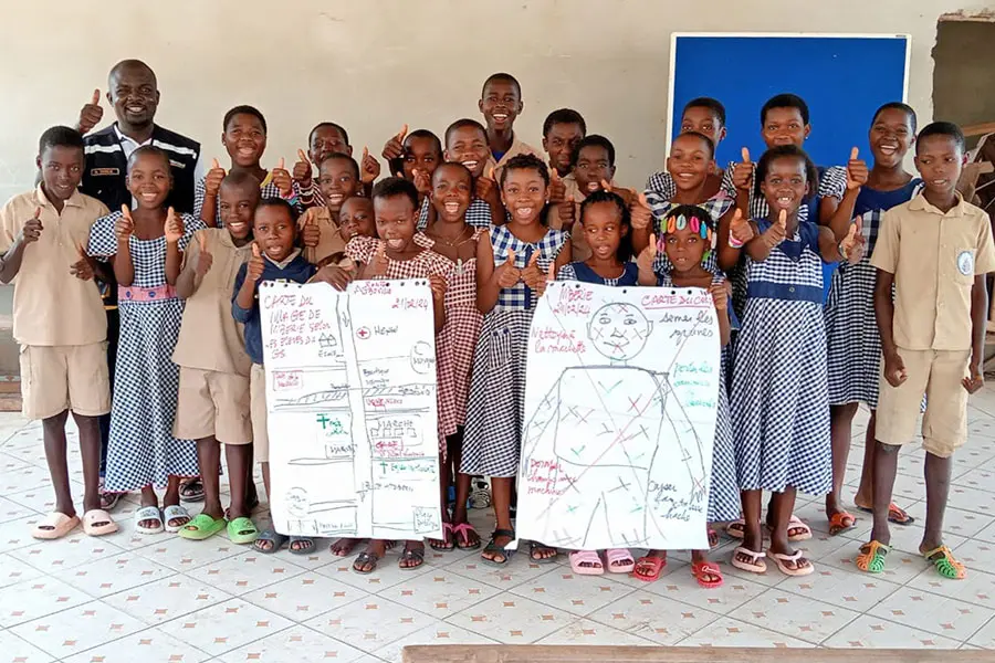 A group of children in matching uniforms pose happily in a classroom, giving thumbs up. They stand around two large drawings on a poster/