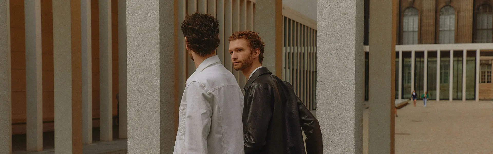 Two men are walking past tall, geometric columns in a modern, urban setting. One wears a white denim jacket and the other a black leather jacket, engaged in conversation. A bicycle is leaning against a column nearby, while the background features more architecture and several people in the distance. The mood is casual and stylish.