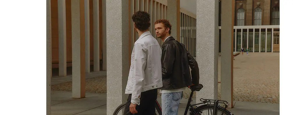 Two men are walking past tall, geometric columns in a modern, urban setting. One wears a white denim jacket and the other a black leather jacket, engaged in conversation. A bicycle is leaning against a column nearby, while the background features more architecture and several people in the distance. The mood is casual and stylish.