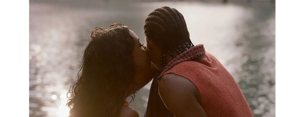 A couple sits on a stone ledge beside a shimmering body of water, sharing a tender kiss. The woman wears a colourful, flowing top, while the man has a textured, orange sleeveless garment. The scene conveys intimacy and warmth, set against a tranquil backdrop.