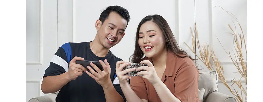 A man and a woman sit together on a light-coloured sofa, smiling as they play games on their smartphones. The woman has long, wavy hair and wears a brown shirt, while the man, with short dark hair, is in a black t-shirt with blue accents. Light wooden panels are visible in the background, adding a relaxed atmosphere.