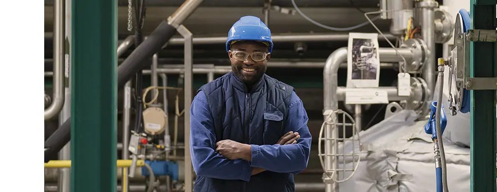 A worker in a blue helmet and glasses stands confidently with arms crossed in an industrial setting, surrounded by machinery and pipes. He smiles, indicating a positive mood.