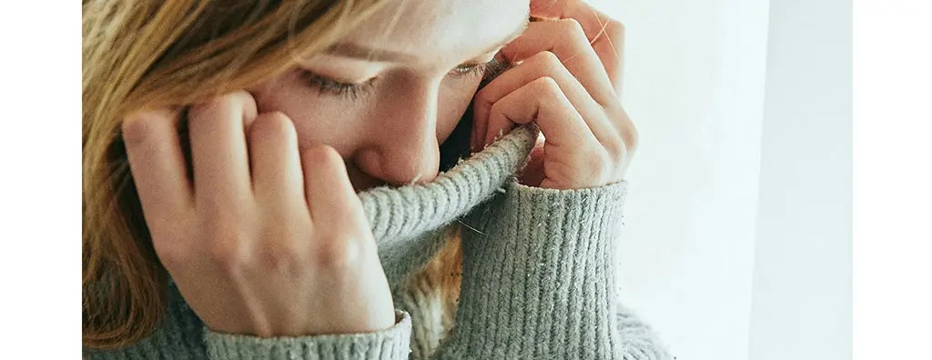 A close-up of a person with long, blonde hair, gently pulling the collar of a grey knitted sweater over their mouth. The soft lighting creates a contemplative mood, emphasising their thoughtful expression.