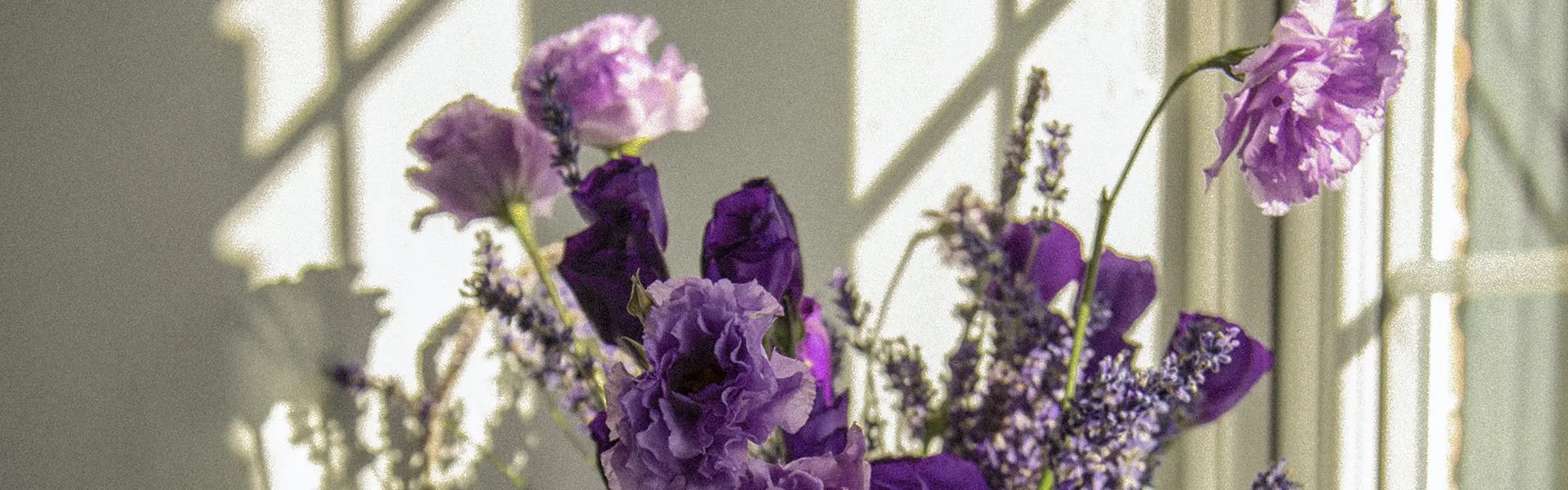 A pale blue vase holds a bouquet of purple flowers, including lilacs and carnations, on a sunlit windowsill. Soft shadows from the window panes create a warm, inviting atmosphere.