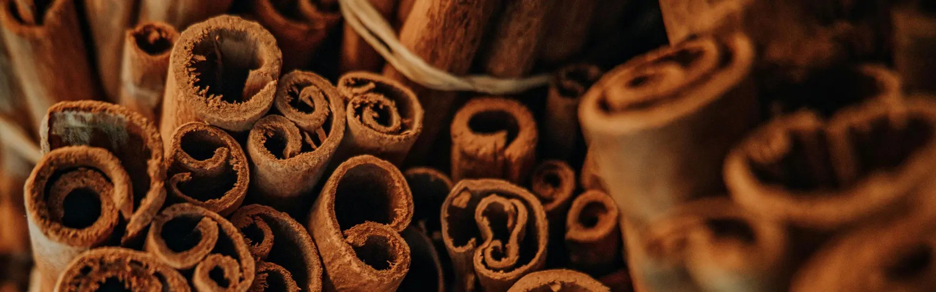 A close-up of rolled cinnamon sticks stacked together, showcasing their spiral shapes and rich brown tones. The image conveys an earthy and warm mood, suggesting a connection to cooking or baking.