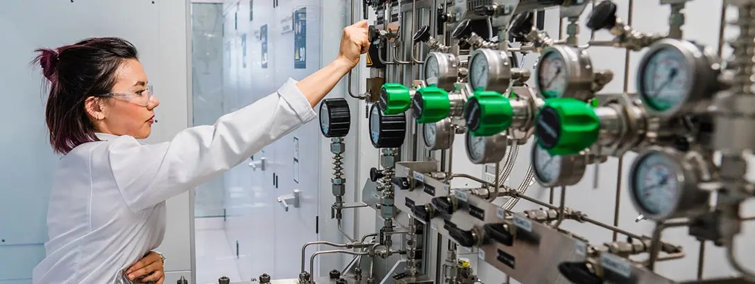A laboratory technician in a white coat adjusts equipment in a modern lab. The workspace features shiny metal machinery, green gauges, and a bright, clean environment, conveying a sense of precision and focus.