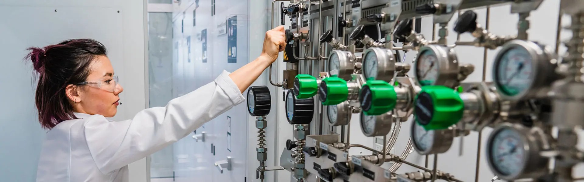 A laboratory technician in a white coat adjusts equipment in a modern lab. The workspace features shiny metal machinery, green gauges, and a bright, clean environment, conveying a sense of precision and focus.