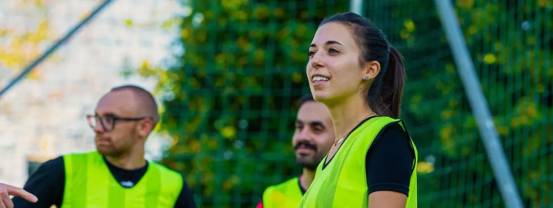 Three individuals participate in a football training session, wearing bright yellow training vests. A woman smiles and engages with the group, set against a green backdrop of trees and goalposts.