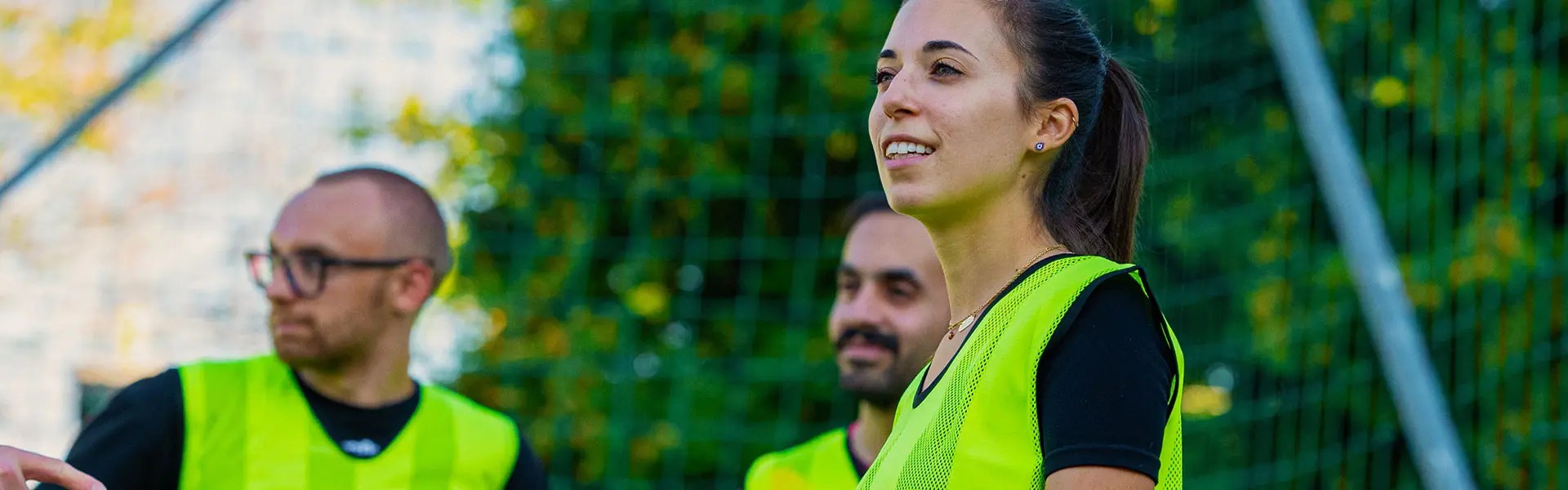 Three individuals participate in a football training session, wearing bright yellow training vests. A woman smiles and engages with the group, set against a green backdrop of trees and goalposts.