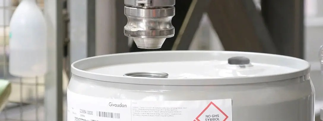 A silver robotic arm is positioned above a grey barrel with a white lid, ready to dispense liquid. The label on the barrel features safety information, and a spray bottle is visible in the background.