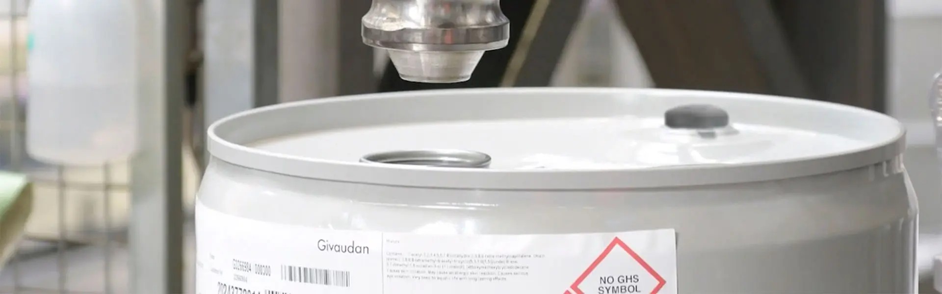 A silver robotic arm is positioned above a grey barrel with a white lid, ready to dispense liquid. The label on the barrel features safety information, and a spray bottle is visible in the background.