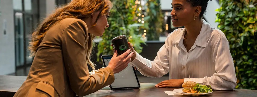 Two women are seated at a wooden table in a green, vibrant setting. One woman, in a beige blazer, hands a Myromi device to the other, who wears a white striped blouse. A laptop and a plate of food are visible on the table. The atmosphere appears friendly and engaged.