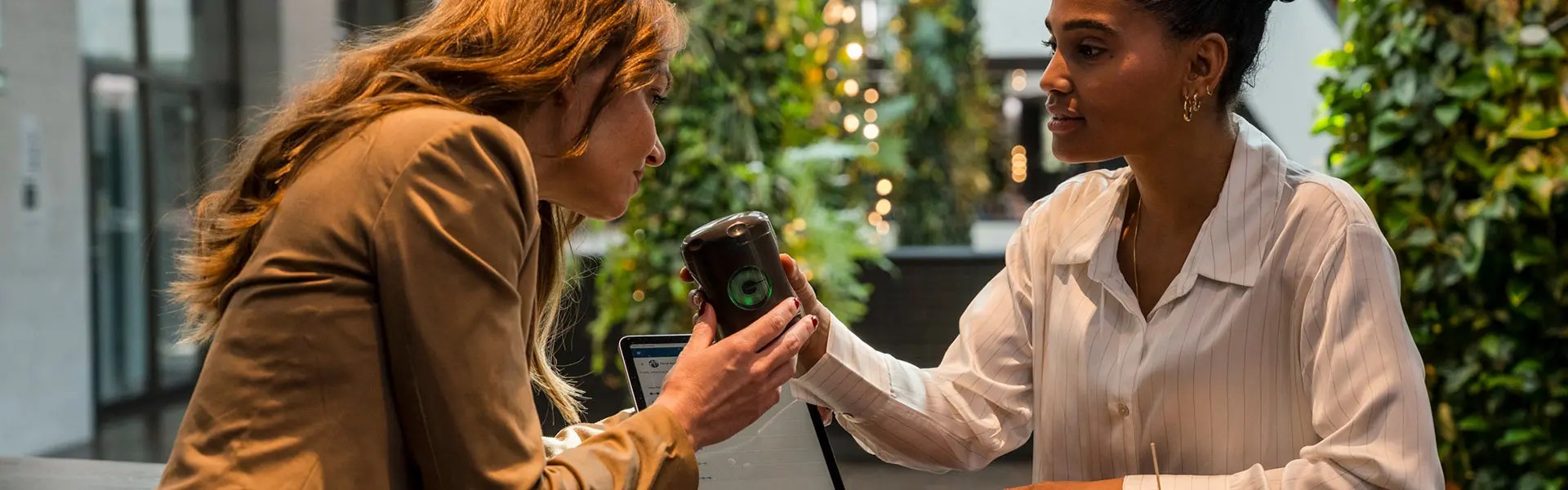 Two women are seated at a wooden table in a green, vibrant setting. One woman, in a beige blazer, hands a drink to the other, who wears a white striped blouse. A laptop and a plate of food are visible on the table. The atmosphere appears friendly and engaged.