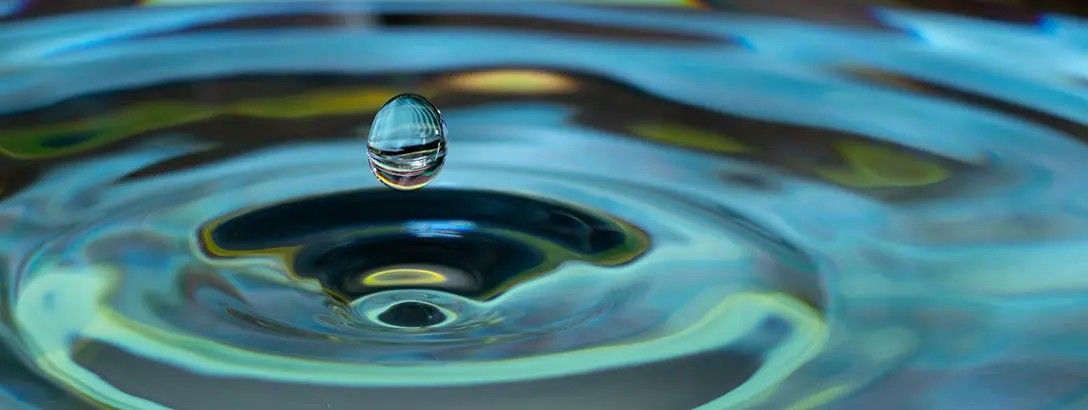 A single water droplet hovers above the surface, creating circular ripples. The background features shimmering blue and green hues, evoking a tranquil mood.