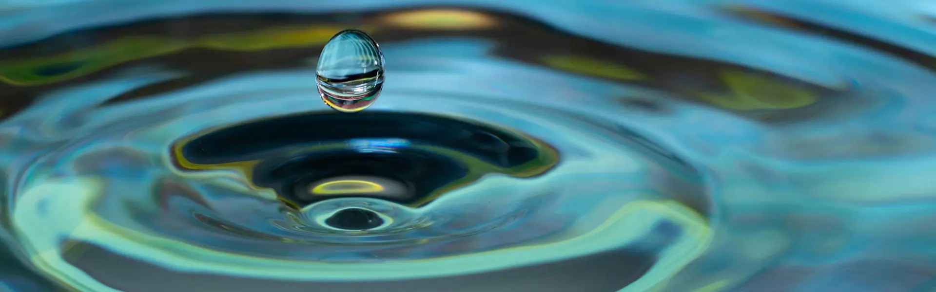 A single water droplet hovers above the surface, creating circular ripples. The background features shimmering blue and green hues, evoking a tranquil mood.