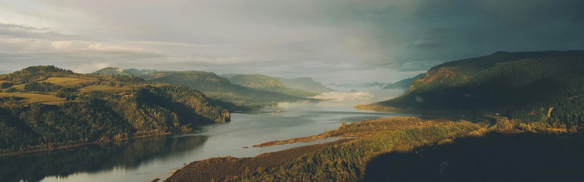 Lush green hills rise on either side of a calm river, reflecting shades of blue and grey from a cloudy sky. Soft sunlight breaks through, creating a serene atmosphere.