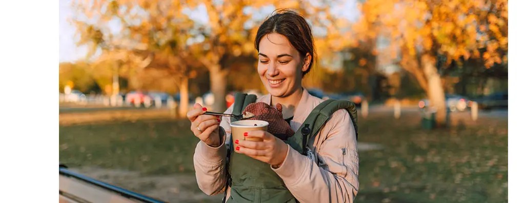 A smiling woman with a green baby carrier holds a cup of food with a spoon in a park surrounded by golden autumn leaves. The setting is warm and cheerful.