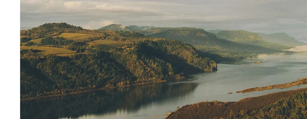 A sweeping view of a river winding through lush, green hills under a dramatic sky. Dark storm clouds contrast with patches of blue, creating a moody atmosphere.