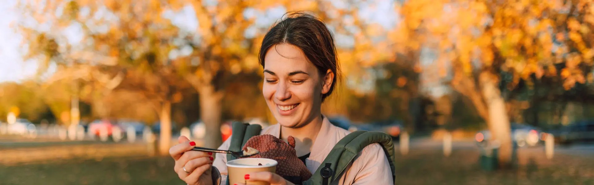 A smiling woman with a green baby carrier holds a cup of food with a spoon in a park surrounded by golden autumn leaves. The setting is warm and cheerful.