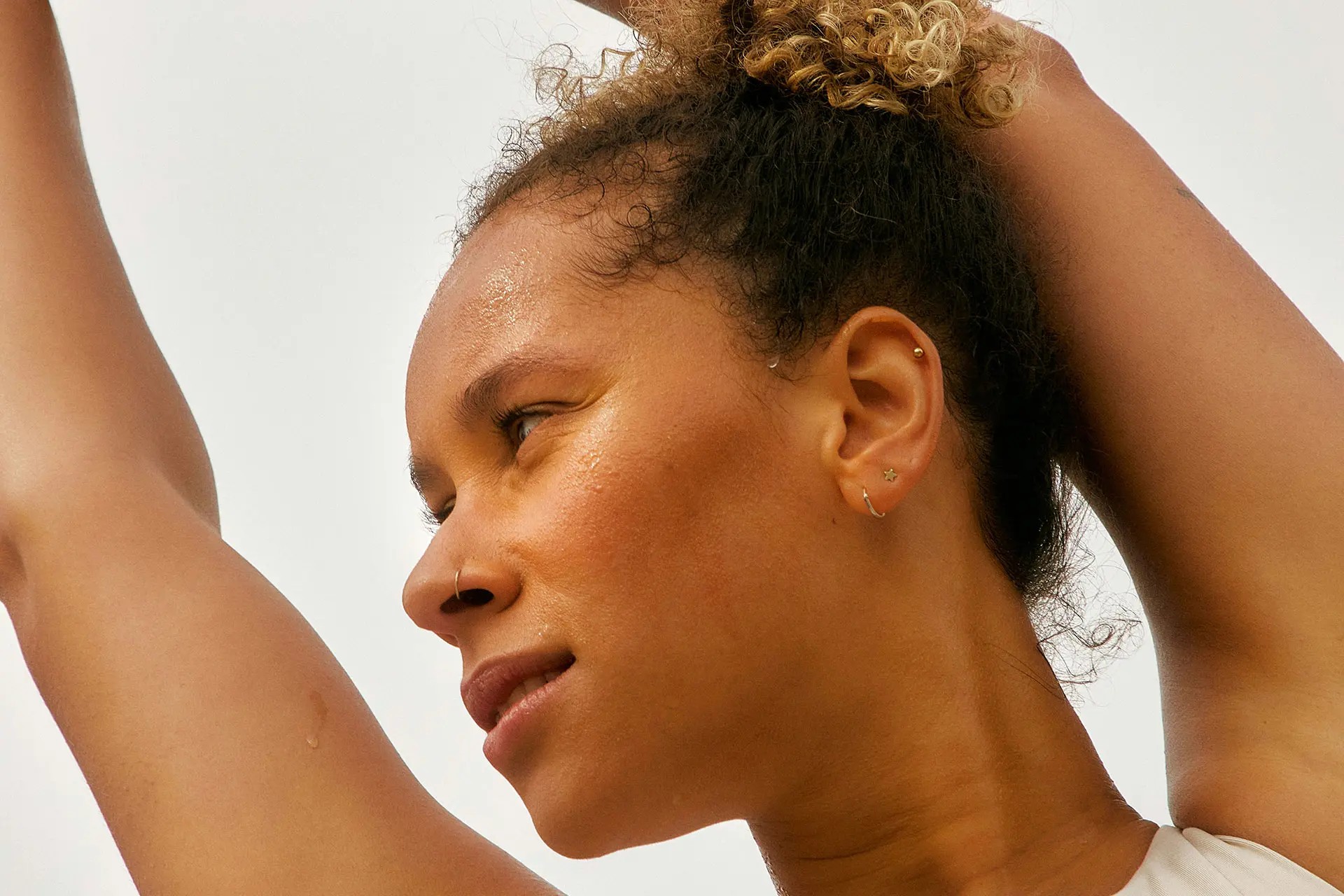 A woman with curly hair gazes upwards, her arms raised above her head. She has a confident expression, with glistening skin suggesting active engagement. The background is softly blurred, enhancing the focus on her face and features.