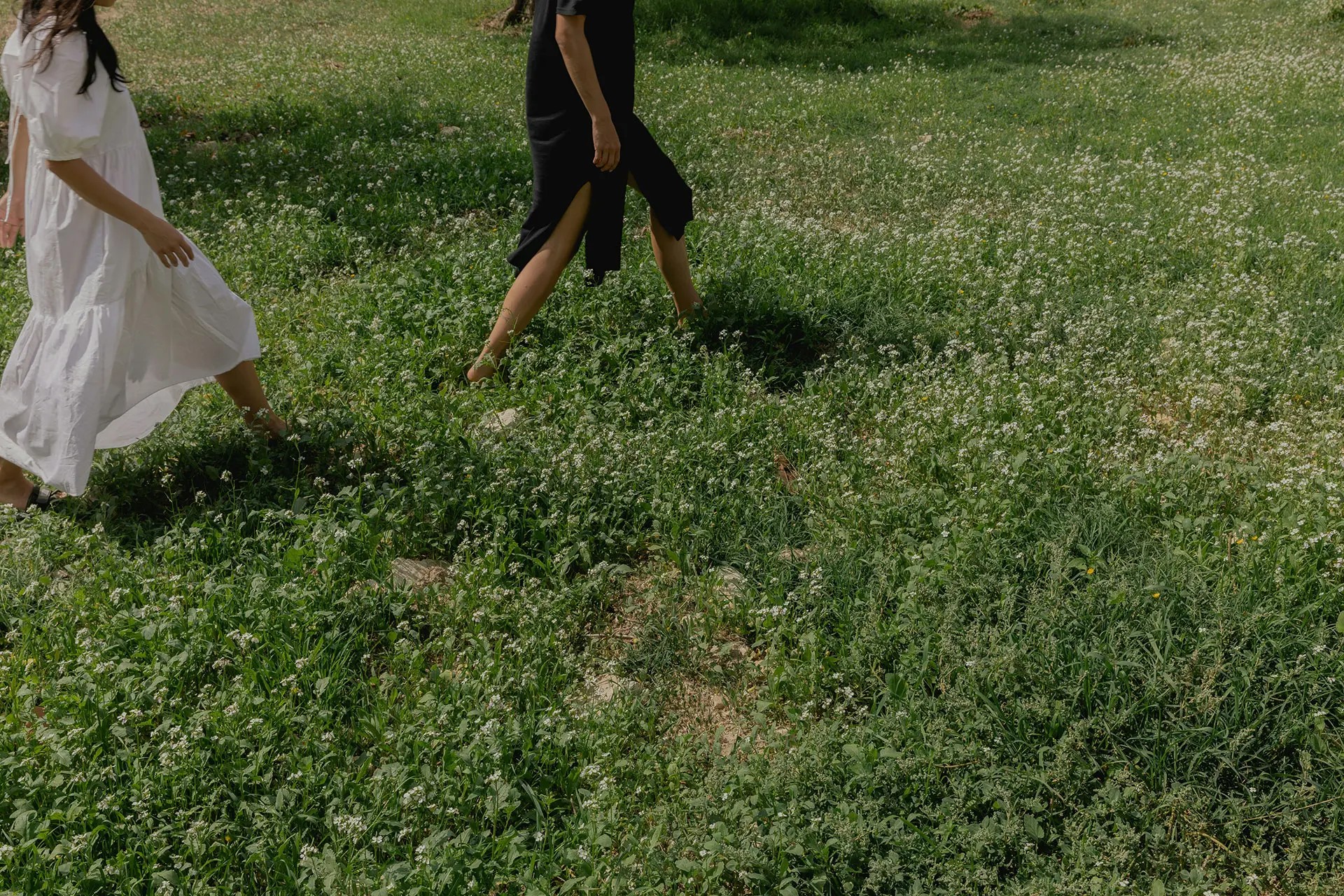 Two people walk barefoot through a lush green meadow filled with white flowers. One wears a flowing white dress, while the other is in a black outfit, both captured mid-step in a serene outdoor setting.